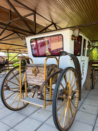 An antique white carriage with gold trim and spoked wheels is parked under a wooden roof in the city of Plastunivska, Russia. The carriage has a wooden chest on its rear, a step for passengers, and two lamps on its exterior. The ground is paved with square tiles. In the background, other carriages and trees are visible.