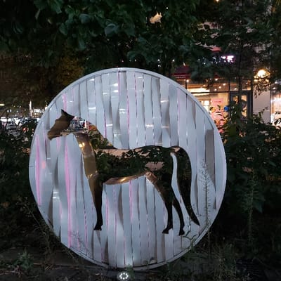 The image displays an outdoor art installation at night in 김포시, South Korea. The central subject is a large, circular sculpture, approximately 1.5 to 2 meters in diameter, composed of vertical white, possibly metallic or plastic, strips. A cutout silhouette of a horse in profile, facing left, is integrated into the center of this circular structure. The horse cutout appears reflective, showing distorted reflections of green foliage and ambient light. Green trees and dense foliage surround and partially frame the artwork. A small, multi-LED ground-level spotlight illuminates the base of the sculpture from the foreground. In the background, across what appears to be a street or open area, commercial buildings with illuminated windows and various light sources are visible. A distinct red sign with white text reading "THE PLACE" is prominently discernible on one of the background buildings, indicating the specific location within 김포시. No individuals are present in the scene.