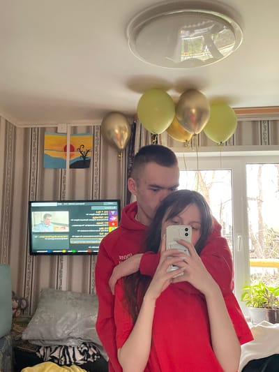 A couple is taking a selfie in a room. The woman is holding the phone, her face partially obscured by it, and she's wearing a red hoodie. The man is beside her, partially obscured, with short hair. The room has a window in the background, revealing outdoor foliage. To the left, there's a wall with a circular light fixture and several balloons (gold and light green). Below the couple, a television screen displays content. To the left of the TV hangs a painting. The location is Ulukulevo, Russia.