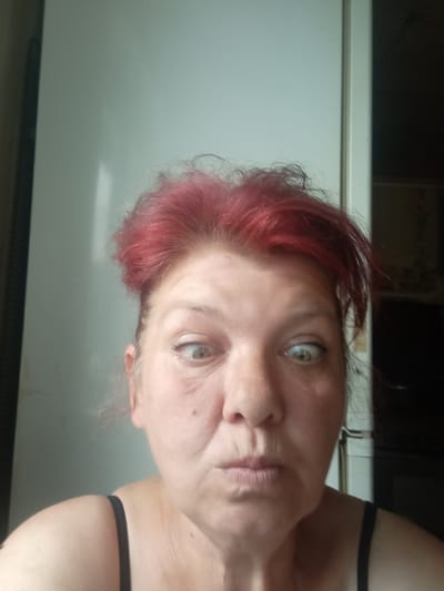 A selfie of a woman with bright red hair, light-colored eyes, and her mouth puckered. She is wearing a black strap top. The background is a wall, possibly a refrigerator, and a dark doorway. The scene appears to be indoors, in what could be a home kitchen. The image does not provide enough information to determine its location in Дукорскі сельскі Савет, Belarus.
