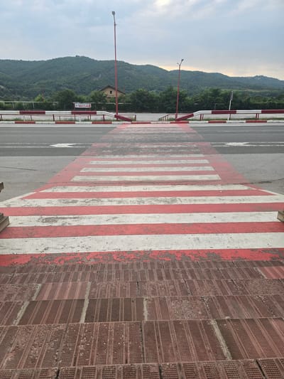 The image depicts a crosswalk in the city of Zlatna Panega, Bulgaria. The crosswalk, painted with red and white stripes, extends towards the viewer from a road surface. In the foreground, a set of brown, textured steps leads up to the crosswalk. The road features traffic lanes and a red and white striped barrier. Beyond the road, a line of trees and a distant, tree-covered mountain range are visible under a cloudy sky. A house with a red roof is situated on the mountainside. Streetlights with red poles are present on either side of the road. A banner with text in Cyrillic is affixed to a fence behind the barrier. The scene is static and appears to be an outdoor public space.