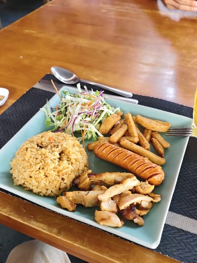 An overhead shot shows a meal on a teal plate, situated on a dark grey placemat on a wooden table. The meal consists of a mound of fried rice, a large grilled sausage, a portion of what appears to be grilled chicken, French fries, and a side salad with shredded cabbage, carrots, and purple onion. A spoon rests on the placemat above the plate, and a fork is positioned to the right of the plate. A glass of yellow liquid, likely a beverage, is partially visible on the far right edge of the image. The wooden table shows a warm, polished finish. The scene appears to be in a restaurant, likely in Phan Rang – Tháp Chàm, Vietnam, given the context.