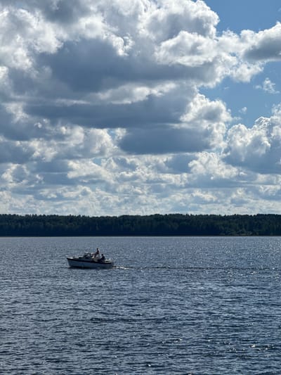 The image captures a vast outdoor scene over a body of water, identified as Lake Saimaa in Lappeenranta, Finland. The sky, occupying the upper two-thirds of the frame, is characterized by numerous dense cumulus and stratocumulus clouds, primarily in shades of grey and white, with intermittent patches of clear blue sky visible, notably towards the right side. A dark green, dense treeline stretches across the mid-ground, delineating the horizon between the sky and the water. The lower third of the image shows the dark blue water of the lake, featuring a textured surface with ripples and abundant small, bright glints of sunlight reflecting off the water. A small, light-colored motorboat with a dark-colored lower hull stripe is positioned in the mid-left section of the water, actively moving from left to right, leaving a faint wake. Two individuals are discernible on the boat: one person is standing towards the stern, and another is seated or standing near the forward cabin section. The overall lighting suggests a bright, partly cloudy day.