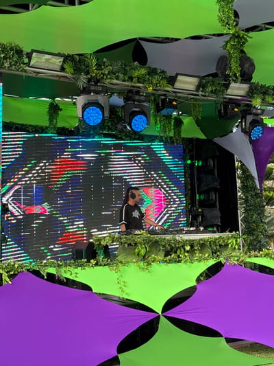 The image depicts an outdoor music stage setup in Noumea, France. A male individual, identified as a DJ, stands behind a white console. He has dark skin, dreadlocks, and wears a black t-shirt with white shoulder stripes and headphones. His right hand is positioned on the console's equipment. Behind him, a large rectangular LED screen displays a digital pattern of horizontal and vertical lines in green, blue, red, and white. Above the screen, a metal truss structure is fitted with multiple stage lights, including two circular moving head lights emitting blue illumination and several rectangular floodlights. Artificial green foliage and vines are integrated throughout the stage, wrapped around the trussing, bordering the screen, and adorning the console. Overhead, a canopy of lime green and grey fabric panels is stretched. To the right of the LED screen, a stack of dark-colored line array speakers is visible. The foreground shows additional lime green and purple fabric panels. The scene is illuminated by natural light.