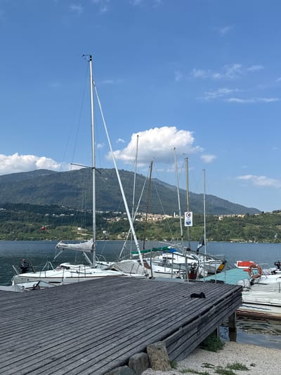 The image depicts a sunlit scene at a marina in Calceranica al Lago, Italy. A wooden dock, constructed from weathered grey planks, occupies the foreground, angled to the right. Several sailboats are moored alongside the dock, their white hulls and tall masts dominating the middle ground. The lake water is a deep blue, reflecting the clear sky. In the background, a verdant, tree-covered mountain range rises, dotted with small buildings, suggesting a town nestled on its slopes. The sky is a bright blue with scattered white clouds. A black object, possibly a sleeping cat, is positioned on the wooden dock in the lower right quadrant of the image. A life preserver ring is visible on one of the boats. The overall impression is one of a peaceful, picturesque day at the lake.
