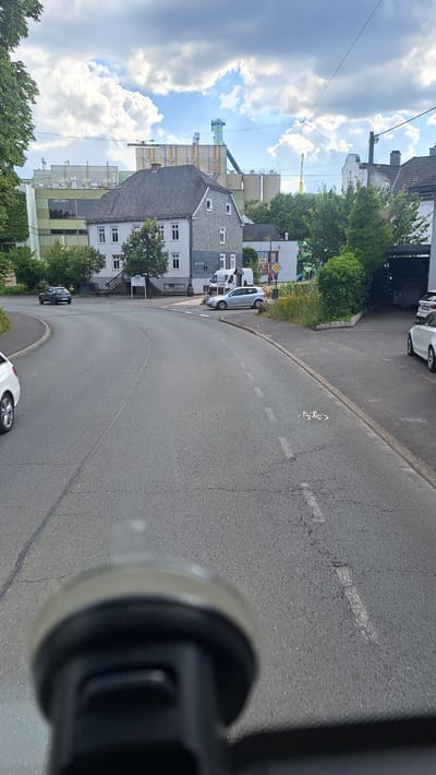 This image captures a street scene in Kreuztal, Germany, on a cloudy day. In the foreground, the edge of a vehicle's interior, likely a dashboard mount or similar apparatus, is visible, slightly out of focus. The street itself is asphalt with white lane markings, including a dashed line and a symbol of a bicycle indicating a bike lane.
The midground shows a curved road leading towards a junction. Several cars are present: a dark SUV is parked or driving on the left side, a silver sedan is parked on the right side of the junction, and the rear of a white car is visible on the far left. A white van is parked near a building.
In the background, a large industrial complex with prominent silos and elevated structures dominates the skyline. Adjacent to the industrial buildings are residential or commercial structures with grey tiled roofs. Green trees and foliage are interspersed throughout the scene, particularly on the left and right sides. The sky is filled with a mix of white and grey clouds.