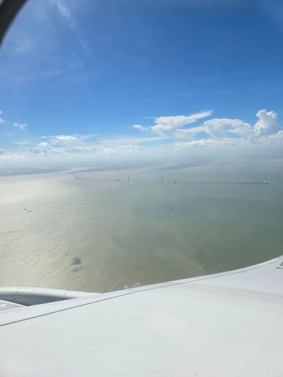 This aerial photograph, taken from an airplane window, captures a panoramic view of the Shenzhen-Zhongshan Bridge spanning across the Pearl River Estuary. The vast expanse of the murky green water is illuminated by sunlight, creating a shimmering effect on its surface. A long, continuous bridge structure stretches across the water, featuring multiple sections and support towers. Several ships and boats are visible on the water, navigating the waterway. In the distance, a hazy shoreline with landmasses and structures can be discerned. The sky above is a brilliant blue, dotted with scattered white clouds, some of which are wispy and others are more voluminous. The lower portion of the image shows the white wing of the airplane, with visible rivets and panel lines, suggesting the perspective is from inside a commercial aircraft. The overall scene depicts a vast aquatic landscape with a significant man-made infrastructure, characteristic of the bustling port city of Shenzhen.
