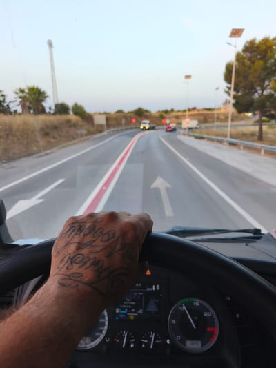 The image is a first-person perspective from the driver's seat of a vehicle, likely a truck, as indicated by the size of the steering wheel and the dashboard instruments. The driver's left hand, which has intricate black tattoos on it, is resting on the steering wheel. The tattoos appear to be script-style lettering. The vehicle is on a road with a red divider line. The road appears to be in a rural or suburban area, with dry grass and sparse trees on the sides. In the background, there are streetlights, one of which has a solar panel. Several vehicles are visible on the road ahead, including a white truck and two cars. The sky is a pale blue, suggesting it is either early morning or late evening. The dashboard displays various readings, including speed and other vehicle information. Based on visual cues such as the vegetation, road type, and the presence of streetlights with solar panels, this scene is consistent with locations found in or around Manises, Spain, particularly along its outskirts or connecting roads.