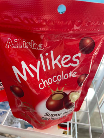 The image displays a close-up of a red, resealable plastic bag for "Ailisha Mylikes chocolate". The bag features a white font for the brand and product name, with the word "chocolate" in a slightly smaller font below "Mylikes". There are visual depictions of chocolate candies on the bag, some whole and one broken to reveal a creamy filling. Text indicating "Super delicious" and "Silky melts in your mouth" is visible. The bag is hanging or placed within a white wire shelving unit, commonly found in stores. The background is a blurred blue, suggesting an indoor environment. There are no people visible in the image. The location of San Jose del Monte, Philippines is not depicted or implied by any elements in the image.