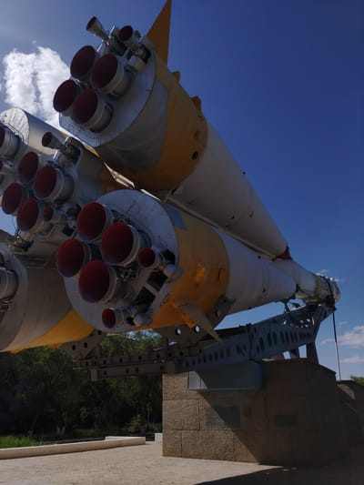 This is a low-angle, full shot of a large rocket on display against a clear blue sky. The rocket is primarily white and yellow, with multiple engine nozzles visible at the bottom, which are red on the inside. The rocket is mounted on a complex, grey metal support structure that rests on a textured, stone-like foundation. The foundation appears to be part of a monument or display area. In the background, trees and foliage are visible, suggesting an outdoor setting. The scene is likely in Baikonur, Kazakhstan, a city known for its space launch facilities.