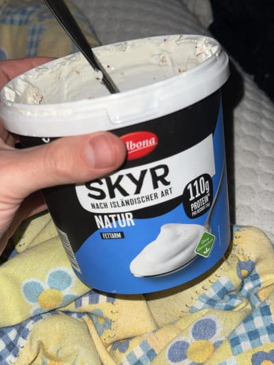 A partially eaten container of Albona Skyr yogurt (variety "Natur Fettarm," indicating "natural low-fat") is being held. A spoon is inside the container. The yogurt container is white with a black and blue label. The text "SKYR NACH ISLÄNDISCHER ART" is prominently displayed on the label. The text "110g PROTEIN" is also visible. The container is being held over a patterned fabric, with yellow and blue flowers and checks. The location is likely Rafz, Switzerland, given Albona is a Swiss brand and the label is in German.