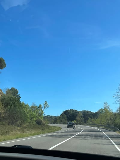 A motorcycle is riding on a two-lane road with a solid white line dividing the lanes. The road curves to the right. The road is bordered by trees and a metal guardrail. The sky is blue and mostly clear. The location is Maçanet de la Selva, Spain.