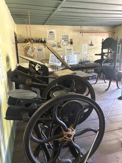 This image depicts an indoor scene likely within a historical printing shop or museum exhibit. The primary subjects are large, vintage printing presses with intricate metalwork, including large flywheels. Several posters and flyers are attached to a wall in the background, some of which are legible. Visible text includes titles like "TOWAGE RIVER ST. LAWRENCE," "MRS. MUMPHRIES BEAUTY BEAST," "GRAND TRUNK RAILWAY," and "SIOO REWARD!! THE MURDERER." There are also notices regarding "Teachers," "HOOL HOUSE," and meeting dates. The setting appears to be a rustic interior with wooden floors and a grey corrugated metal wall on the left. The lighting is consistent with an indoor setting, suggesting daytime. There are no people visible in the image, and the printing presses appear to be static, implying a display rather than active operation. The overall impression is one of a preserved historical space showcasing the technology of early printing.