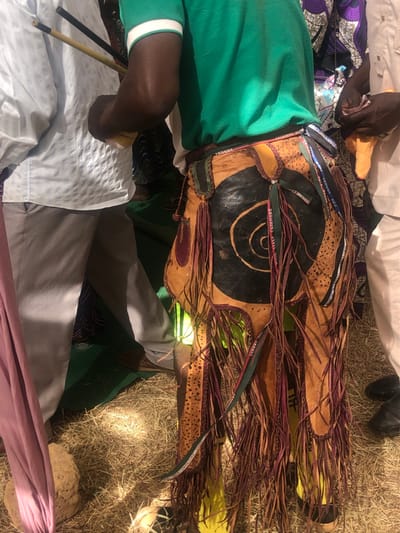 The image captures an outdoor gathering in Kerang, Nigeria, centered around a person wearing or presenting a richly adorned leather garment or accessory. The setting is a dry, sunlit area covered with light brown straw or grass.
The most prominent subject is an elaborate piece of light brown leather, possibly animal hide, featuring a large black painted circle in the center with concentric lighter rings. The leather is decorated with numerous strips of fabric or ribbon in various colors (green, purple, red), and long, reddish-brown leather fringe. Some sections of the leather also have embossed patterns resembling animal spots. Several ribbons incorporated into the design bear visible text, including "WOOL ITALY CASHMERE," "SUPER VINE," "LUXURY WO...", and "GURKHA."
A person, visible on the left side of the frame, is wearing a green long-sleeved shirt with a white and green striped cuff and light-colored trousers. This individual's arm and hand are seen holding two thin sticks, one black and one yellow/light brown. Other attendees are partially visible around the central figure, dressed in various fabrics including white, light blue, purple patterned, and pink clothing. Dark shoes are also discernible on the ground.
The bright daylight and distinct shadows indicate a daytime event. The scene suggests a cultural or ceremonial gathering, possibly involving a traditional performance or display of craftsmanship, as indicated by the intricate and distinct nature of the leather object and the sticks held by the individual. The use of branded ribbons hints at the incorporation of contemporary or repurposed materials into traditional artistry.