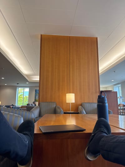 A first-person perspective photograph captures an interior lounge or common area within a building in Waltham, United States. In the immediate foreground, two feet are prominently displayed, propped up, resting on or near a polished wooden table that spans the lower half of the frame. The left foot wears a black suede-like slipper or boot, while the right foot wears a dark suede mule or clog with a visible light-colored sole, paired with dark striped socks. Dark trousers cover both legs.
On the wooden table, a closed black laptop or tablet (possibly a Lenovo brand, indicated by a faint logo) is positioned to the left, and a dark blue or black insulated water bottle with a flip-top lid is to the right.
Beyond the table, a tall, vertical wooden partition, composed of light-brown wooden panels with a prominent vertical grain, dominates the center of the image. To the right of this partition, another polished wooden surface with chairs is visible in the background. To the left of the partition, a seating area is arranged with multiple striped armchairs (grey and beige) and small side tables. One small table lamp with a white square shade and a slender silver base illuminates the area between two armchairs adjacent to the wooden partition.
In the far left background, a large window reveals a vibrant green outdoor scene, suggesting trees and possibly a body of water or distant structures. Natural light permeates from this window. A blurred figure wearing headphones is partially visible seated in an armchair in this section. The ceiling is white with integrated linear lighting fixtures and square acoustic panels, providing bright ambient illumination. The overall scene suggests a relaxed study or waiting area.