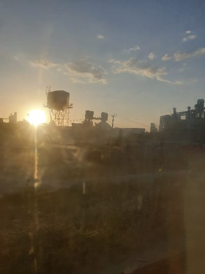 A silhouetted industrial landscape meets the vibrant sunset sky. Water towers and skeletal frameworks stand against a blend of blue and yellow hues, with a radiant sun peeking through. Power lines stretch horizontally between structures. The scene evokes a sense of industrial dominance juxtaposed against natural beauty, captured through a tinted window.