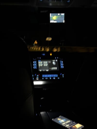 The image shows the interior of a vehicle at night. The infotainment system is visible on the dashboard, displaying various information. In the back, a passenger entertainment screen is visible, angled towards the viewer. Outside the window is the blur of the urban environment at night, with lights of buildings visible. The foreground is dominated by the rear seat of the car. It is impossible to verify the image's location is in Kuchak, Armenia.