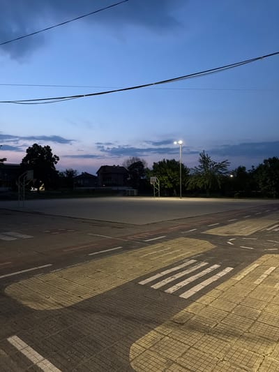 The image depicts an outdoor urban environment in the city of Saedinenie, Bulgaria, captured during twilight or early evening. The scene is devoid of human figures or active vehicles.

The upper portion of the frame is occupied by a twilight sky, transitioning from a deep blue overhead to a lighter purplish hue near the horizon, with scattered dark grey clouds. Two prominent dark utility cables or power lines traverse the upper third of the image horizontally.

The midground features an outdoor paved area, consistent with a multi-purpose court or playground. On the left side of this court, a metal basketball hoop with a white backboard and a green support structure is visible. A second, partially obscured basketball hoop is present on the right side of the court. A tall, silver-colored streetlamp with a dual light fixture is centrally positioned within the court, emitting bright white light that casts shadows across the ground.

The foreground consists of a patterned paved surface, primarily dark grey square tiles, overlaid with various road markings. A distinct white-striped crosswalk is visible in the lower right. Yellow-striped areas and dashed white lines, simulating road features, are also present, suggesting a recreational area possibly designed for children's traffic education. Shadows, likely cast by the illuminated streetlamp and possibly unseen structures, are visible across this patterned pavement.

In the background, behind the sports court, a dense line of dark-silhouetted trees forms a barrier. Partially visible through and above the trees on the left side are several residential buildings, appearing as two-story houses, characterized by dark, muted tones consistent with the low light conditions.
