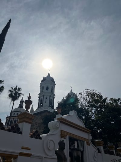 An upward angle shot showcases a white church tower with a cross atop against a bright sun and cloudy sky. Ornate architectural details are visible on surrounding structures, including sculpted figures. Trees and palm trees frame the composition. The image has a slightly tilted perspective and washed out colors due to the sun's brightness.
