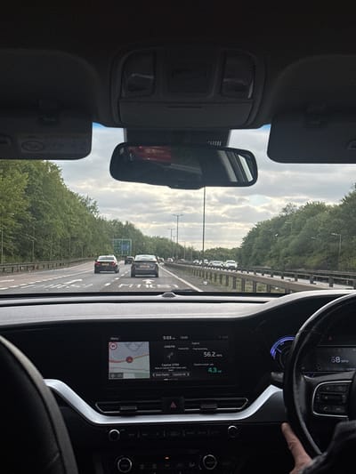 The image depicts a car interior, specifically the driver's perspective. The car is on a highway, with other vehicles visible ahead. The dashboard displays information such as speed (68 mph), fuel economy (56.2 mpg), and a navigation map indicating a speed limit of 80. The radio is tuned to Capital XTRA. The environment appears to be a wooded area alongside the highway, under a cloudy sky. The location is likely within Uttlesford, United Kingdom, based on the "Capital UK" radio station selection. A person's hand is visible on the steering wheel.