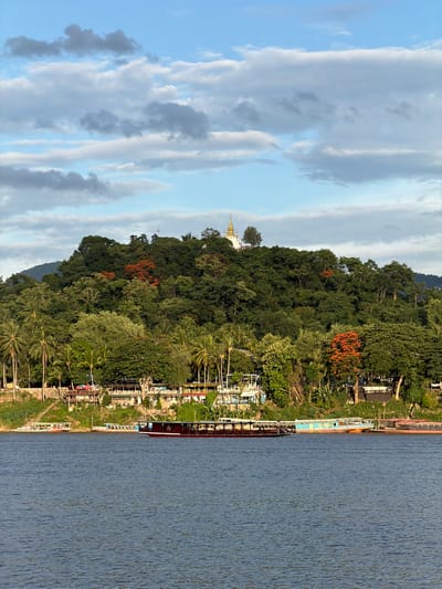 A scenic view of the Mekong River in Xieng Men cultural village, Laos, showcases a lush green hillside topped by a gilded temple. Several boats, including a large red and white vessel, are docked along the riverbank. The sky is partly cloudy, with streaks of white and gray against a bright blue backdrop. The dense foliage on the hillside includes palm trees and other tropical vegetation, with a cluster of vibrant orange flowers adding a splash of color. Buildings, likely shops and restaurants, are visible along the waterfront. The water of the river is calm and appears slightly rippled.