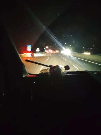 The image displays a forensic, matter-of-fact scene captured at night from inside a vehicle on a multi-lane highway, identified as being in the vicinity of Three Mile Cross, United Kingdom. The dark vehicle dashboard is visible at the bottom of the frame, with a windscreen wiper arm extending from the lower left. A clear plastic bottle, appearing partially full and consistent with a Coca-Cola product due to its shape and a visible red label section, rests horizontally on the dashboard. Reflections and minor streaking are present on the windshield. Beyond the windshield, a multi-lane highway recedes into the darkness. Numerous red taillights from vehicles ahead indicate moderate to heavy traffic flow in the same direction. Intense white glare and lens flare emanate from approaching or adjacent vehicle headlights in the midground and background, illuminating sections of the road. White solid and dashed lane markings are visible. On the left side of the highway, a white crash barrier parallels the road. Two distinct road signs are present: a blue rectangular sign with an indistinguishable graphic, and a smaller white triangular warning sign. The sky above the highway is completely dark, confirming nighttime conditions.