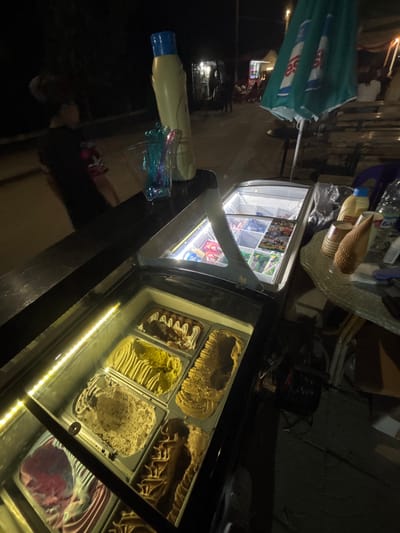 The image depicts an outdoor vendor stand at night, described as being in Kostievo, Bulgaria. In the immediate foreground, a black counter surface is visible, upon which stands a tall, light-yellow plastic bottle with a blue cap and a clear container holding multiple colorful plastic scoops. Directly below the counter is a glass-fronted display freezer containing six large metal trays of various ice cream or gelato flavors, including chocolate, vanilla, a speckled light brown, a ridged yellow, and a pink/red swirl. An LED strip illuminates the interior of this freezer. To the right and slightly behind, a second, smaller glass-fronted freezer displays numerous pre-packaged frozen items, likely ice cream bars or popsicles, arranged in baskets.
To the right of the freezers, a round, translucent table with a textured base holds a stack of brown paper cups, a large textured conical object, and a clear plastic bottle with a partially visible Cyrillic label ("БА"). A dark green market umbrella, partially folded, stands behind the table, displaying a white logo with two stylized 'e's. A dark purple plastic chair is partially visible near the table. On the ground beneath the table, a black electrical cable reel is present. In the background to the left, a person wearing a dark shirt with a colorful shoulder design is partially visible and blurred. The overall background features a dimly lit street or market area with other distant stalls or establishments marked by artificial lights, and several indistinct figures of people suggesting a populated environment.
