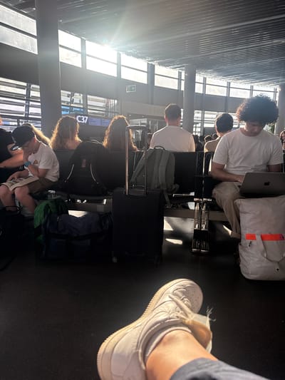 A sunlit waiting area in Schwechat, Austria, at gate C37, features a row of people occupying metal and black mesh chairs. Most are occupied with electronic devices or books. Luggage, including roller suitcases and backpacks, is adjacent to the chairs. A white Nike Air Force 1 shoe is prominent in the foreground. The time displayed on the gate information screen is 16:50 for a Ryanair flight to Dubrovnik.