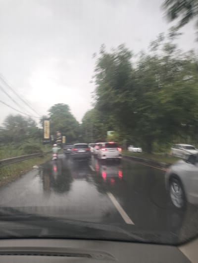 The image captures a blurry scene of a traffic jam on a wet road, likely during the day under overcast skies, indicating rainy weather. The perspective is from inside a vehicle, showing the dashboard in the foreground. Several cars are visible on the road, with their brake lights illuminated, suggesting slow-moving or stopped traffic. One car in the foreground has its headlights on, and a motorcycle with a rider wearing a light-colored helmet is also present, positioned behind some of the cars. The road is reflective, indicating the presence of puddles or a generally wet surface.
On the left side of the road, there is a grassy embankment bordered by a metal guardrail. Further back, lush green trees line the road, creating a natural, somewhat dense foliage. In the background, there are two tall, rectangular billboards with yellow and dark lettering. One billboard is slightly taller and more prominent, with what appears to be text in a vertical orientation. The overall atmosphere is subdued, consistent with a cloudy or rainy day. The motion blur suggests the vehicles are either moving slowly or the photo was taken while in motion. The location is identified as Santan, Indonesia.