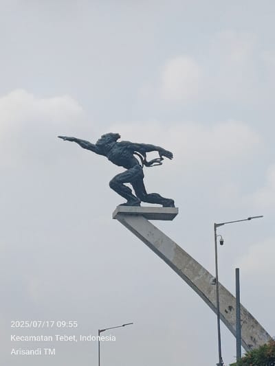 The image depicts a large, dark-patinated bronze statue of a male figure in a dynamic, forward-striding pose, mounted on a light-colored, upward-sloping concrete pedestal. The figure's left arm is extended straight forward, and its right arm is bent with the hand near the chest, with flowing elements trailing behind its back. The background consists of an overcast sky with scattered light gray clouds. To the right of the statue's base stands a tall metal street light pole with a downward-angled lamp and a spherical CCTV camera mounted below it. A second street light pole is partially visible in the lower left, and green foliage is present in the bottom right corner. The image includes a timestamp of 2025/07/17 09:55 and is located in Kecamatan Tebet, Special Capital Region of Jakarta, Indonesia. No individuals are visible.