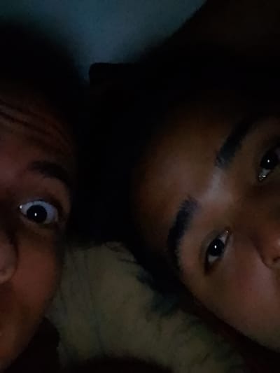 Two individuals are featured in a very low-light, close-up shot, likely taken indoors at night in El Espinal, Venezuela. The image is tightly cropped on their faces, suggesting a selfie or a very intimate photograph.
On the left side of the frame, one person's face is largely visible, looking directly at the camera with wide, alert eyes and a slightly surprised or playful expression. The whites of their eyes are prominent against their dark skin tone. The upper part of their head is cut off by the frame.
On the right, the second person's face is partially visible, primarily showing one eye, an eyebrow, and parts of their forehead and nose, also with a dark skin tone. This individual appears to be looking slightly upwards or to the side, with a calmer or more contemplative expression than the first. Their hair is dark and blends into the deep shadows of the background.
The overall scene is characterized by extreme darkness, with subtle hints of blue and green tones in the background, possibly from faint ambient light reflecting off surfaces or bedding. The lack of distinct features or objects in the background reinforces the focus on the subjects' faces. No visible text is present in the image.