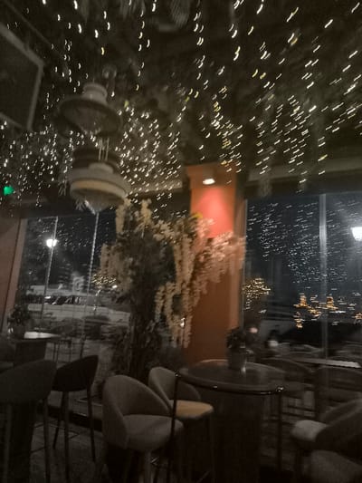 This image captures the inviting interior of an establishment, likely a restaurant, bar, or cafe, at night. The ceiling is a canopy of numerous small, warm-toned string lights, creating a sparkling, magical effect, intermingled with dark foliage. Two large, ornate hanging fixtures, possibly decorative lights or sculptures, are visible in the upper center.

In the mid-ground, a prominent, tall decorative plant with abundant light-colored, drooping flowers stands alongside a reddish-orange pillar that is softly illuminated by an upward-facing light. The foreground features several modern, upholstered chairs or stools with light-colored seats and dark frames, arranged around a round, dark-toned table on which a small potted plant rests.

A large glass window dominates the background, revealing the outside world at night. Through the window, streaks of distant artificial lights, including streetlights and what appear to be urban or possibly coastal lights, are visible, creating a blurred, dynamic backdrop. No individuals are present in the scene. The overall atmosphere is cozy and warm, achieved through the abundant use of decorative lighting against the dark setting. The image has a soft blur, suggesting low light conditions or camera motion. There is no clearly readable text visible. The view through the window hints at an urban or coastal location, consistent with Palaio Faliro, Greece.