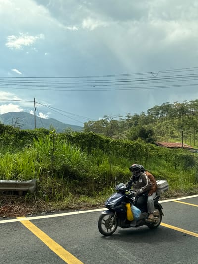 Two individuals are riding a dark blue motorcycle with a white storage box attached to the rear. The person in front is wearing a helmet with "Cafe PH" printed on it, a patterned face mask, a brown jacket, and light gray pants. The person behind is wearing a brown jacket and light gray pants. The motorcycle is on an asphalt road with a yellow line. To the left of the road is a metal guardrail bordering lush green vegetation and tall grasses. In the background, a hillside is covered in dense green trees and foliage, with a glimpse of a building with a rusty tin roof. Power lines are visible strung across the sky, supported by a utility pole. The sky is partly cloudy with a mix of white clouds and a lighter blue hue. The scene appears to be on the outskirts of Tu Poh, Vietnam, suggesting a rural or semi-rural setting.