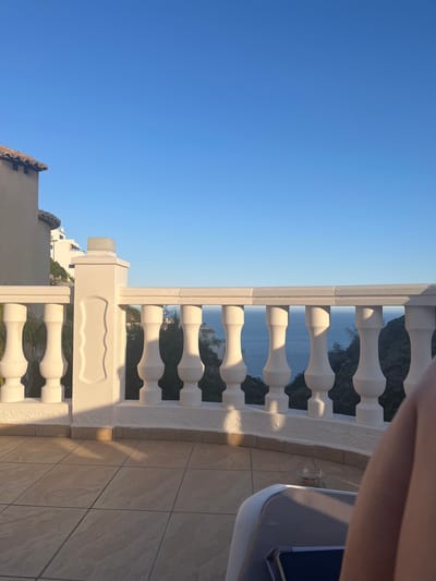The image shows a view from a balcony in El Poble Nou de Benitatxell/Benitachell, Spain. The balcony features a white balustrade with classic baluster shapes. Behind the balustrade, a glimpse of the ocean and vegetation is visible. A clear blue sky fills the upper portion of the frame. The balcony floor is tiled in light brown with subtle patterns. A section of a white outdoor lounge chair is seen in the upper right corner. A mobile phone is placed on the lounge chair. The image appears to be rotated 90 degrees counter-clockwise. In the lower left, a building with terracotta roof tiles is seen. The balustrade partially obstructs the view of surrounding buildings nestled among the trees.