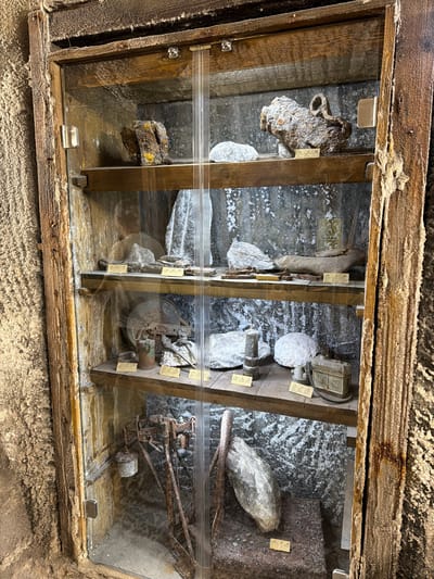 The image shows a wooden display case, likely in the Turda Salt Mine, located in Boj-Cătun, Romania. Inside the case, there are several old mining artifacts and rock samples arranged in three separate compartments. The artifacts include rusted metal objects like pipes, a fan, and what appears to be a portion of a mining cart or machinery. There are also stones and what appear to be salt formations. The back of the case, which serves as the background for the artifacts, shows a rough, textured surface that resembles the mine walls. The display case is encased in salt formations.