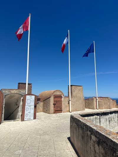 The image shows a view of the Citadel of Saint-Tropez, a historical fortification located in Saint-Tropez, France. Three flags are flying on flagpoles against a clear blue sky. The leftmost flag is red and white with a green emblem. The middle flag is the French tricolor (blue, white, and red). The rightmost flag is the flag of the European Union.

In the foreground, there is a stone-paved area and a low stone wall with weathered and mossy surfaces. To the left, an arched opening leads into a dark interior, with a white sign posted on the wall inside, featuring text and a map. A wooden door with a metal handle and a sign depicting a map of the citadel is ajar to the right of the opening. Behind this, there are tan-colored, rough-textured structures with arched doorways and small windows. A brick chimney is visible on the left. The sea and distant hills can be seen in the background on the right. The scene is illuminated by bright sunlight, casting shadows. There are no people visible in the image.