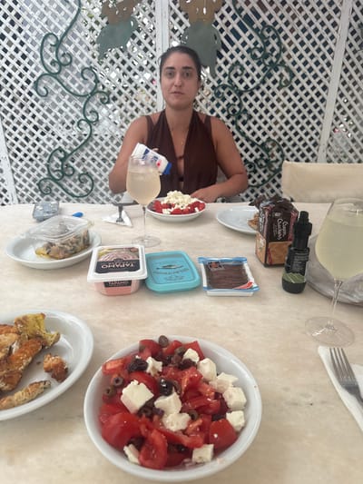 The image depicts a table laden with food and drinks, suggesting a meal in an outdoor setting, likely in Naoussa, Greece. A young woman is partially visible on the left side of the frame, her upper body and face in focus as she looks towards the camera. She is wearing a brown top. The table itself is covered with a light-colored surface, possibly marble.

On the table, various food items are arranged. There's a white bowl filled with a Greek salad, containing chopped tomatoes, cubes of feta cheese, and black olives. Next to it, another white oval dish holds what appears to be breadsticks or croutons. A package of "Cream Crackers" by Papadopoulos is open, and beside it, a small bottle with a black cap. Further to the right, a small blue container labeled "HELLAS MEZE" is visible, alongside another small tub with pinkish contents and Greek writing that includes "ΤΑΡΑΜΟ" and "TARAMA." There are also small anchovy fillets in a plastic package.

A glass of a pale yellow drink, possibly white wine or a cocktail, is positioned on the table, next to a wine glass. A fork rests on a white napkin. The background reveals a white lattice structure, characteristic of outdoor seating areas, with some green decorative elements visible. The overall impression is one of a relaxed meal, possibly lunch or an early evening aperitif, enjoying the Mediterranean atmosphere. The lighting suggests it might be daytime.