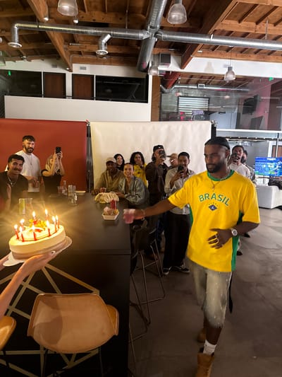 The scene is an indoor celebration, potentially a birthday, in what appears to be a commercial or industrial space converted for event use, possibly in Culver City, United States of America. The architecture features exposed wooden beams and industrial-style ductwork. A diverse group of people are gathered around a long, dark-colored table. Some individuals are holding and using smartphones, seemingly to capture photos or videos of the celebration. A person in a yellow Brazil soccer jersey stands out, possibly the focus of the gathering. Another person presents a lit birthday cake with candles. There are drinks and other items on the table. A backdrop of white and red is visible behind the group. There is also a television in the background.