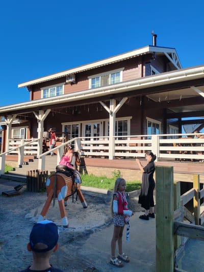 In the city of Lukaši, Russia, a brown, wooden, two-story house with white trim and a porch stands under a clear blue sky. Several people are present on the property. In the foreground, a young boy wearing a blue and black baseball cap faces away from the camera, looking towards a wooden hobby horse with a girl riding it. The girl, wearing a pink dress and with a cast on her left arm, is also looking away from the camera. Behind them, a woman in a black dress stands with her hands clasped together, facing the house. Further back on the porch of the house, another person wearing a hat and carrying a basket walks up the stairs. The ground appears to be dirt or gravel, with some grass patches visible.