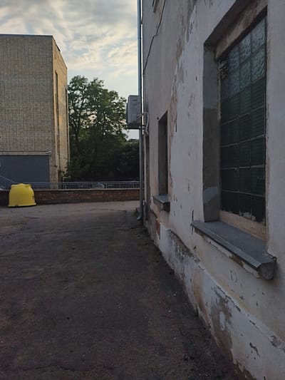 This media file depicts an outdoor scene, likely in Tukums, Latvia. The foreground is dominated by a dark, asphalt-covered ground, possibly a courtyard or parking area. To the left, a tall, light-brown brick building with rows of windows stands under a cloudy sky. In front of this building, a bright yellow, large cylindrical container, possibly a waste bin or storage unit, is positioned near a metal railing and some steps.

The right side of the image features a close-up of an older, weathered building with a pale, peeling facade. This building has several windows, one of which is prominently visible and appears to be made of green, opaque glass blocks. The window frame is weathered, and some of the glass blocks seem damaged. The building's exterior shows signs of age and disrepair, with noticeable cracks and discolored patches on the walls.

In the background, between the two buildings, lush green trees are visible, suggesting a natural element within the urban or industrial setting. The sky above is overcast with a hint of light, implying it could be late afternoon or early morning. There is no discernible activity or event taking place; the scene appears still and possibly abandoned or infrequently used. The overall mood is somewhat somber due to the weathered state of the buildings and the muted lighting. No text is visible in the image.
