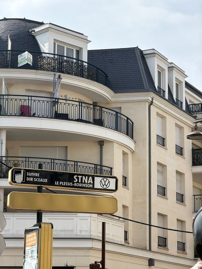 This is an outdoor, eye-level, full-shot photograph of a building and its surroundings in Châtenay-Malabry, France. The building, likely an apartment complex, features a light-colored stone facade with numerous windows framed by white trim. Some windows have black metal grates. A dark, sloped roof with visible tiling covers the upper section of the building. Balconies with black metal railings are visible on the curved sections of the building.
In the foreground, two street signs are mounted on a pole. The left sign is black with white text and a white icon, reading "SUIVRE DIR SCEAUX STNA LE PLESSIS-ROBINSON". The sign to its right is a plain golden rectangle. Below these signs, a smaller sign is attached to the building, displaying text that includes "A VENDE" and "A VENDRE" along with a phone number.
The sky is overcast and appears bright, suggesting daytime. The overall scene conveys an urban setting, possibly in a residential area. There are no people clearly visible in the image, but a pink shopping cart can be seen on one of the balconies. The angle of the photograph is slightly tilted.