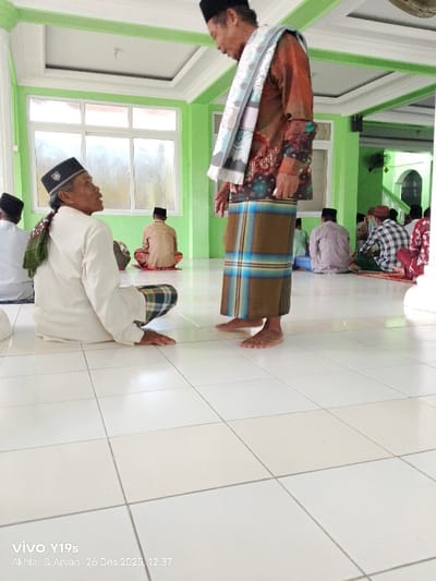Inside a bright, green-trimmed room with tile flooring, a man in a brown sarong, patterned shirt, and shawl stands barefoot, conversing with another man seated on the floor, wearing a white garment and patterned cap. Several people sit cross-legged in the background, some on prayer rugs. Sunlight streams through large windows. A fan is visible on the wall. The date "26 Des 2025, 12.37" is at the bottom left, possibly from a camera overlay.