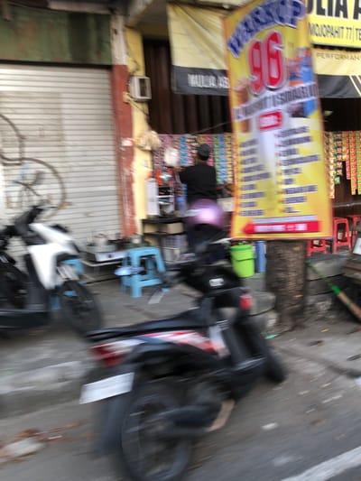 The image depicts a busy street scene in Sidoarjo, East Java. In the foreground, a person wearing a black shirt and cap is partially visible, with a faint purple light emanating from their head, suggesting a camera flash or a similar light source. They appear to be looking down, possibly at a phone or other device.

To the left of the person, a yellow banner is prominently displayed. The banner features large, red numbers "96" and a series of vertical bars of varying heights, along with some text that is difficult to read due to the blur. Below this banner, there is a closed metal shutter door, painted in white, with some green and brown accents on the wall above it.

In the background, there are several objects on the street, including a black motorcycle with its wheels clearly visible, a blue plastic chair or stand, and various other items that are blurred. A rack of colorful goods, possibly snacks or drinks, is visible on a shelf to the left, behind the yellow banner. Red chairs are stacked in the upper right background. The overall impression is one of an urban setting with shops and commercial activity. The lighting suggests it could be daytime. The image is quite blurry, making it challenging to discern fine details.