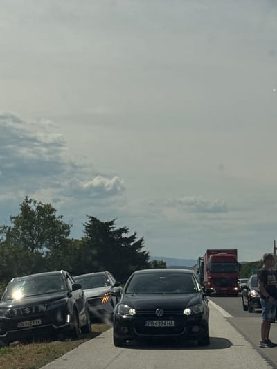 The image depicts a traffic jam on a sunny day, likely in the afternoon, given the bright lighting. The scene is set on a paved road with grassy verges and trees lining the side. The sky is partly cloudy.
Several vehicles are visible, including a black Volkswagen, a white car, and another black car with the license plate "CB 6324 XK". Further back, a red truck is also part of the traffic congestion. On the right side of the road, a person wearing a dark t-shirt and denim shorts is standing, looking at their phone. The person appears to be waiting or observing the traffic.
The overall mood is one of stalled movement, typical of a traffic jam. The image does not convey any specific emotions beyond the passive experience of being stuck in traffic.
Visible text includes the license plate "PB 4994 HA" on the Volkswagen and "CB 6324 XK" on the other black car. The location context provided suggests Shtarkovo, Bulgaria.