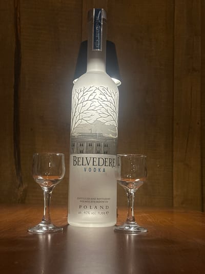 A Belvedere vodka bottle, frosted white with a grey tree silhouette and a depiction of a building, stands on a wooden surface. Two small, clear shot glasses are placed in front of the bottle, one on each side. The background is composed of dark, vertical wooden planks. The lighting is dim, with a spotlight illuminating the bottle and glasses from behind. There are no people present. The location is the city of Lapės, Lithuania.