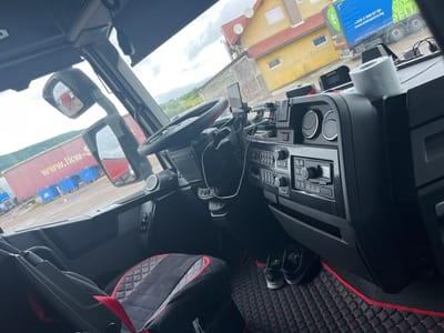 The image offers a wide-angle view from the interior of a truck cab, looking out through the front and side windows. The setting appears to be a parking or staging area for large vehicles.

Inside the cab, the driver's perspective reveals a black and red themed interior. A black steering wheel with red accents is visible on the left, with a smartphone mounted on the dashboard near it, connected by a white charging cable. The dashboard features several controls, including air vents, a radio unit, and various buttons, many set against a textured, carbon-fiber-look panel. A Telepass device and another labeled "GPS TPAR" are on the dashboard. A roll of white toilet paper with a floral pattern rests prominently on the passenger side of the dashboard. From the overhead console, a microphone connected by a coiled cable hangs down. The floor is covered with black, quilted floor mats featuring red stitching and red trim, with a pair of black sneakers placed on the passenger side. The driver's seat, partially visible, has a matching black and red quilted cover.

Looking out, the sky is overcast, and the ground appears damp, suggesting recent rain or generally wet conditions. In the immediate foreground outside the truck, a red trailer is visible, with "www.lkw-s" partially legible on its side. Beyond this, blue dumpsters are lined up. To the right, a distinctive yellow building with a red tile roof and satellite dishes stands, with a covered pile of firewood nearby. Further in the distance, another blue truck trailer is visible, bearing text including "BERGER ecotra," "+359 2 948 07 00," "www.transpress.bg," and partially, "RUST RELIABILITY RELIA." The phone number and website suggest a connection to Bulgaria, while "LKW" (a German acronym for truck) and "Berger Ecotra" indicate broader European trucking references. The location context provided is Petroșnița, Romania.