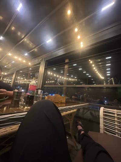 The image depicts an indoor setting at night, possibly a restaurant or cafe, with a prominent glass ceiling and walls. String lights with yellow bulbs are visible along the ceiling, casting a warm glow. In the foreground, a person's legs and feet are visible, wearing black pants and sandals. They are seated at a table, on which a bottle of Coca-Cola and several glasses are placed. The background shows reflections on the glass surfaces, revealing more string lights and the silhouettes of other people seated at tables. The overall atmosphere appears relaxed and social, suggesting a casual evening gathering. The presence of Coca-Cola with its distinctive red branding is a notable detail. The lighting indicates it is nighttime. The context of Qaskeleñ, Kazakhstan, does not provide specific visual cues within the image itself.