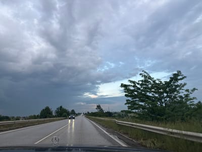 This media file depicts a forward-facing view from a vehicle driving on a wet asphalt road under a cloudy, overcast sky, likely during the late afternoon or early evening. The road appears to be a highway or a major road, indicated by the presence of multiple lanes and a guardrail. Several vehicles are visible on the road ahead, with headlights illuminated. On the left side of the road, there are trees and some distant buildings, suggesting a rural or semi-rural setting. On the right side, there is a dense line of trees and vegetation. The sky is filled with dark, dramatic clouds, with hints of lighter sky visible towards the horizon, suggesting either the approach or departure of rain. The wet road surface and the mood of the sky evoke a sense of impending or recent rainfall. The emblem of a Mercedes-Benz is visible on the hood of the vehicle from which the photo is taken. The location is identified as Ivanyane, Bulgaria. No visible text is present in the image.