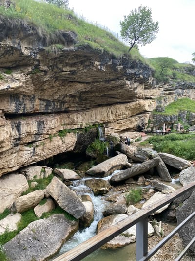 The image depicts a rocky gorge with a stream flowing through it, located in Salta, Russia. Massive, stratified rock formations dominate the scene, with lush green grass and small trees growing on the slopes and along the top of the gorge. A narrow waterfall cascades down the rock face on the left side of the image.
In the mid-ground, a path winds through the rocks, and two people are visible standing on it. Further back, a seating area with umbrellas and tables suggests a café or restaurant. The stream is filled with large, scattered rocks, creating a natural, rugged landscape. A metal railing runs across the foreground, partially obscuring the view of the stream and rocks below. The sky is overcast, indicating a cloudy day. The overall impression is one of a natural, outdoor recreational area.