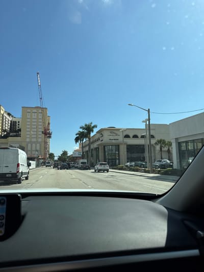 A street view from inside a vehicle, showing traffic on a sunny day. Tall buildings line the road, including a Jaguar Palm Beach dealership. A construction crane is visible near a light-colored building. Palm trees and streetlights are interspersed along the road.